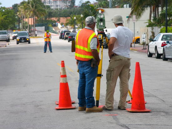 Construction and city workers in safety gear using surveying equipment on a city street with traffic cones, palm trees, and parked cars.