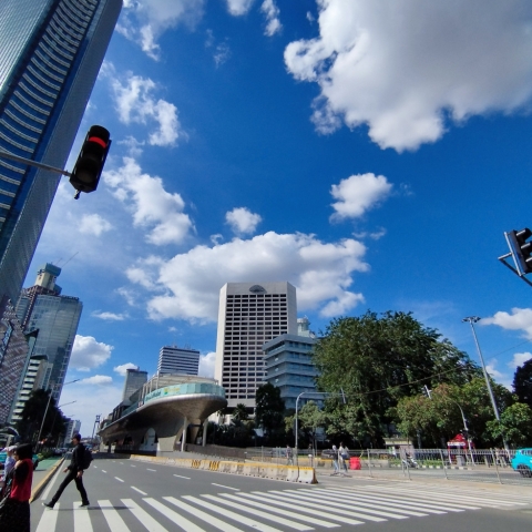 Urban street with zebra crossing, modern glass buildings, elevated transit structure, traffic lights, and pedestrians under a bright blue sky.