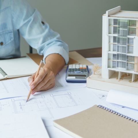 Architect working on building plans with blueprints, calculator, notebook, and residential scale model, illustrating urban economics concepts.