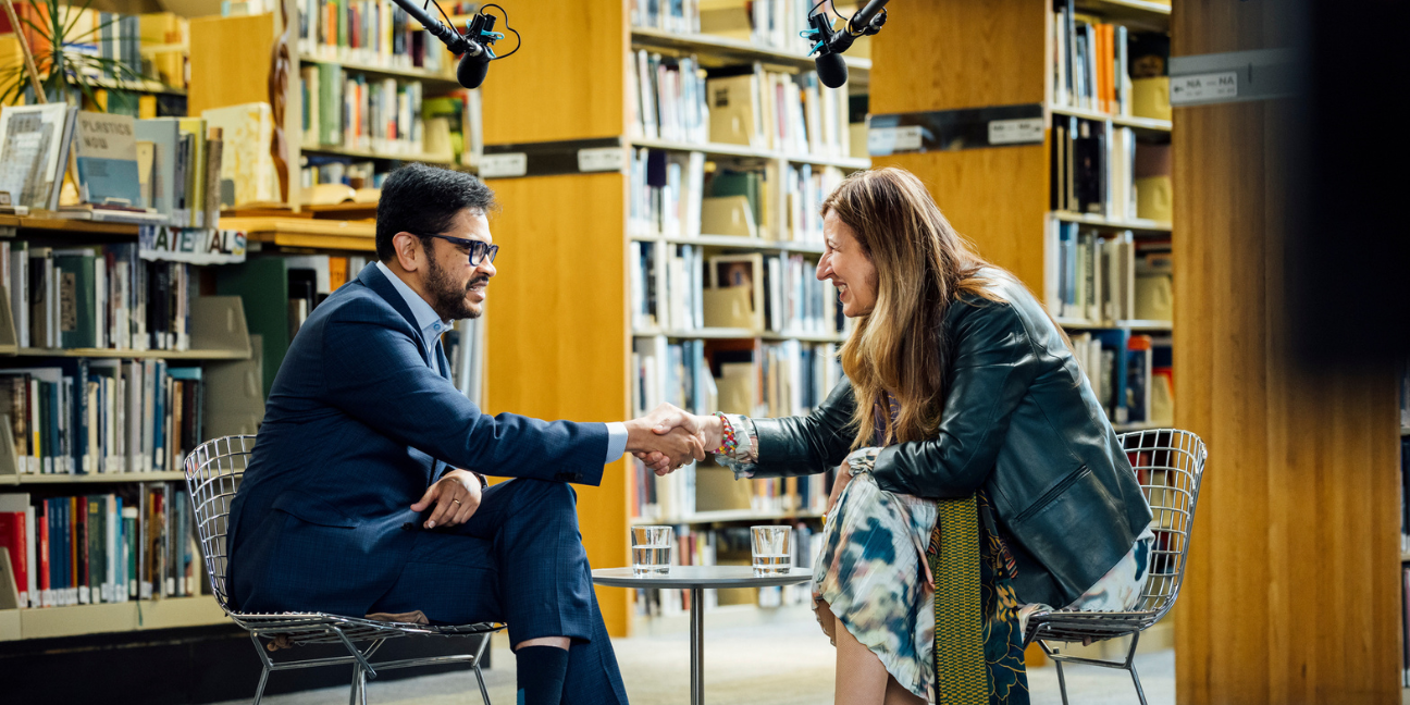 BAC President Mahesh Daas and Benedetta Tagliabue shake hands at the end of a recorded conversation in the BAC library, seated among rows of architecture books.