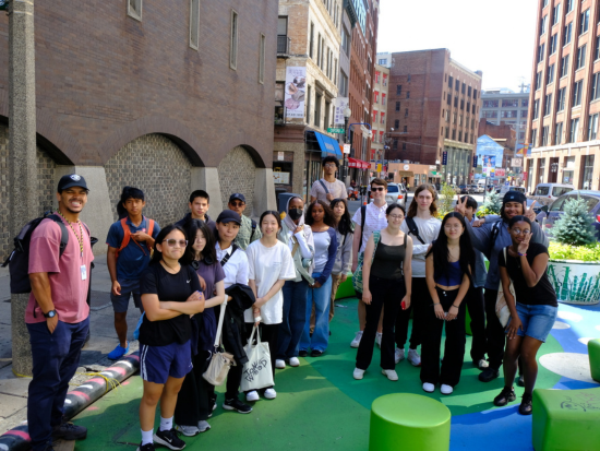 A group of Summer Academy students stands together in a colorful urban plaza during a city field trip, surrounded by tall buildings and bright street art.