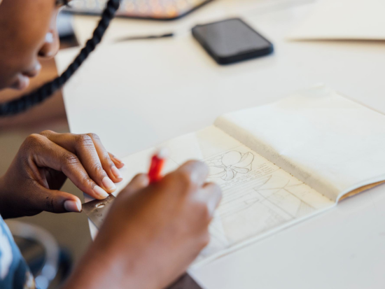 Student sketching an architectural concept in a notebook with a red pencil during BAC Pre-College Urban Design and Planning Studio work session.