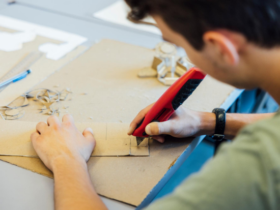 A student using a utility knife to cut cardboard for an architectural model during hands-on work in the BAC Pre-College Urban Design and Planning Studio.