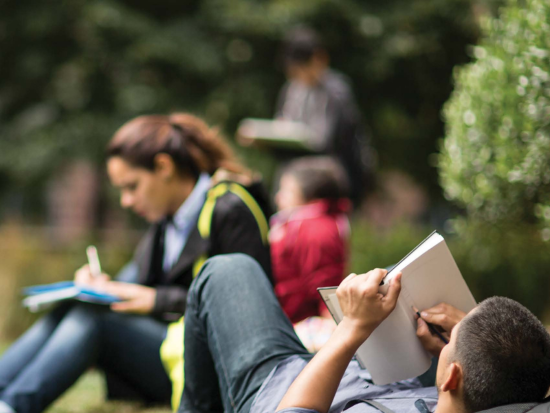 Students work in a notebook while laying on a lawn outside.