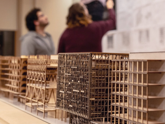 Wooden stick models of buildings (foreground) and students working (in the background).