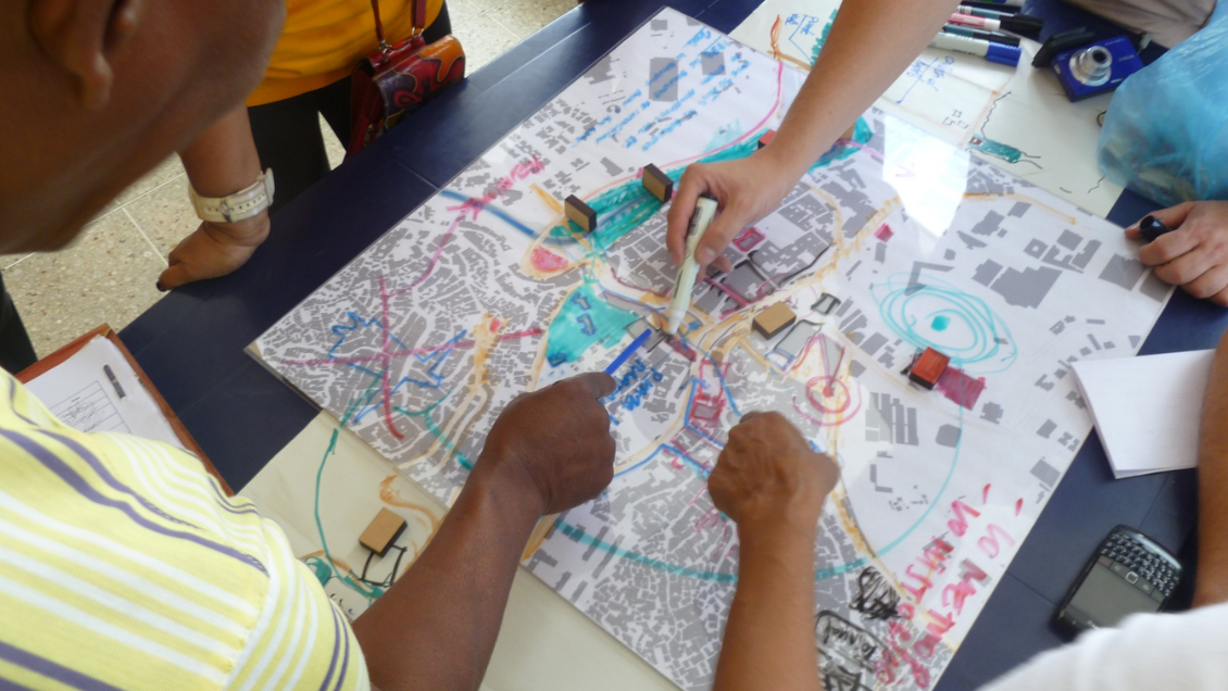 Participants use a map, markers, and blocks during a participatory design workshop in Petare, Caracas (2014), exploring community planning concepts.