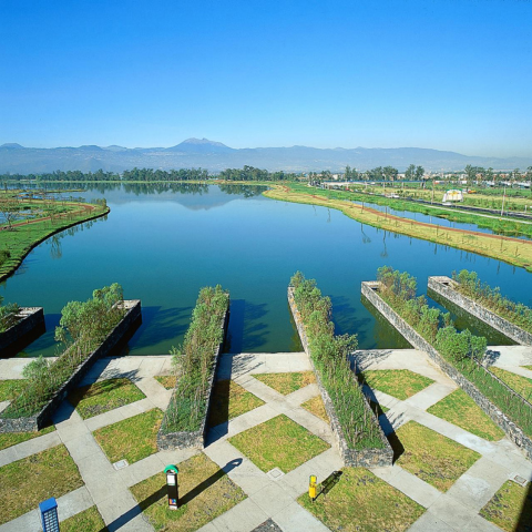 Water flows from a series of stone fountains at the entrance plaza of Xochimilco Ecological Park, framed by wetland vegetation and trees.