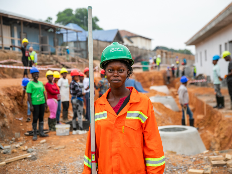 A woman in a bright-orange, construction uniform; a BHI trained Sierra Leonean team member helping in the construction of the Maternal Center of Excellence, a regional facility that will elevate the landscape of maternal care. Over half of the construction workforce are women.