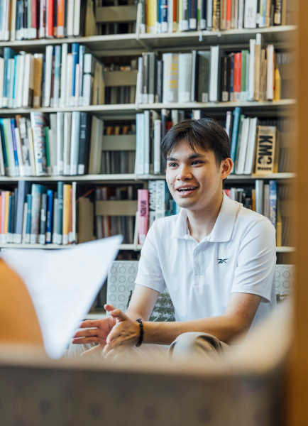 Preston Tran, Summer Academy and UDP Alum, and 2025 Summer Academy teaching assistant, sits in a quiet library space discussing a project, surrounded by tall bookshelves filled with design resources.