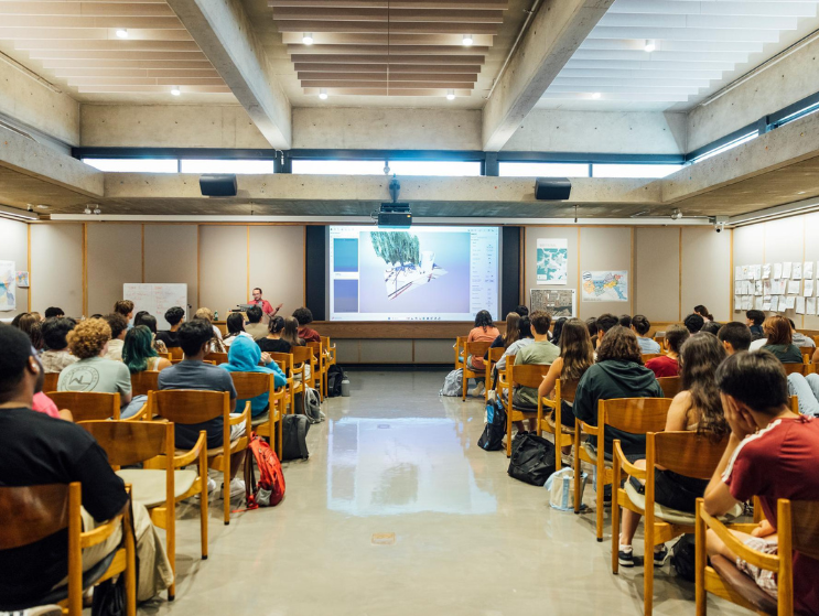 A large group of Summer Academy students sits in the Cascieri Lecture Hall watching a presentation on architectural design projected at the front.