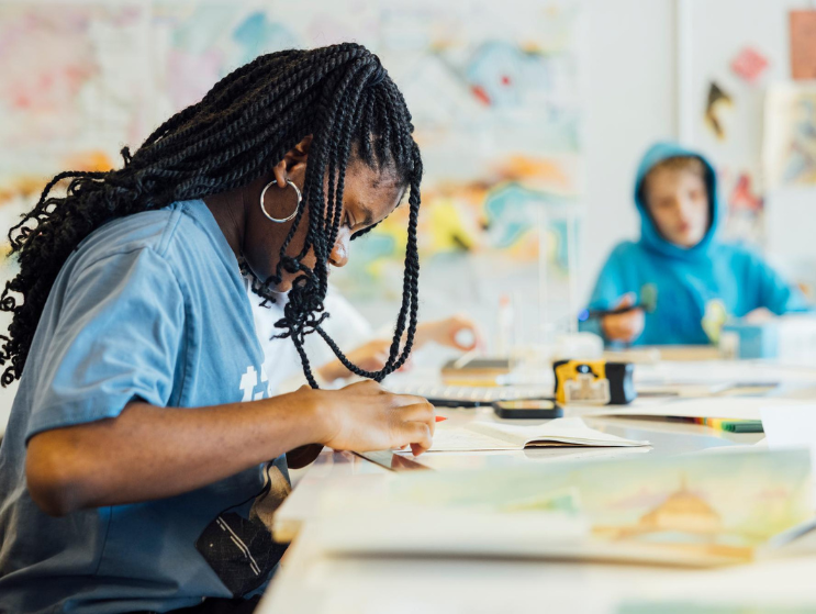 A student focuses on drawing in a bright studio classroom, surrounded by art supplies and colorful design work on the walls.