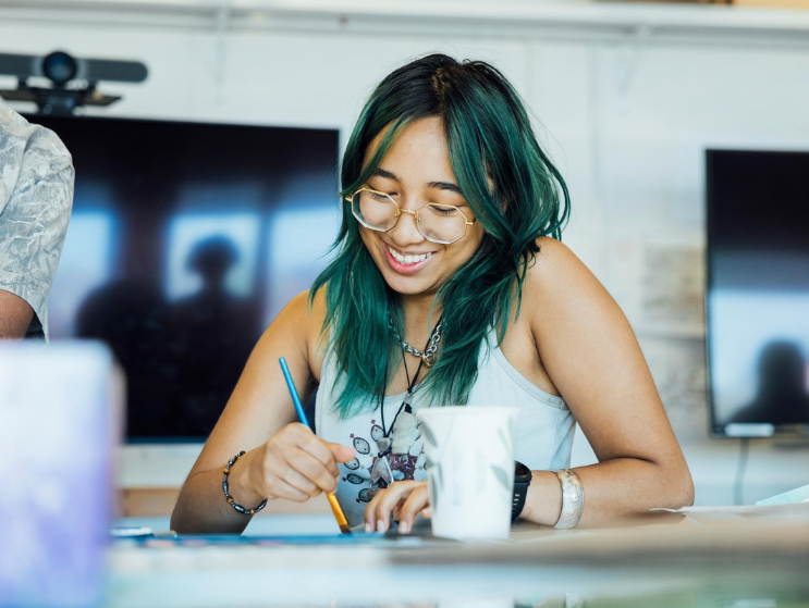 A smiling, summer academy student works on an art project, using watercolors to color a sketch at a studio table.