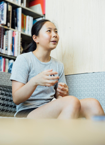 Summer Wu, a Summer Academy student, sits on a lounge chair in a library area, gesturing while speaking during a conversation.