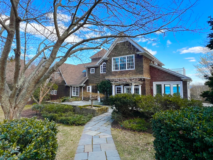 Coastal-style shingle home with stone walkway, landscaped yard, and mature trees under a blue sky in a quiet suburban neighborhood in Jamestown, RI.