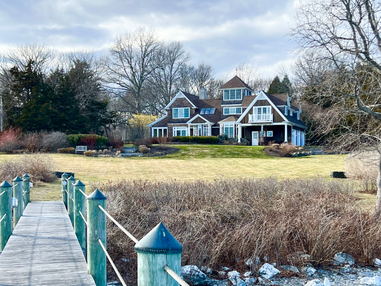 Coastal-style shingle home with stone walkway, landscaped yard, and mature trees under a blue sky in a quiet suburban neighborhood in Jamestown, RI.