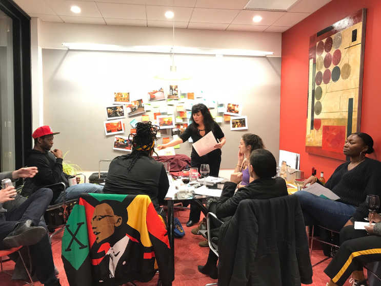 Felice Silverman leads a group gathered around a table for a collaborative design discussion, reflecting the mentorship, dialogue, and community central to design practice.